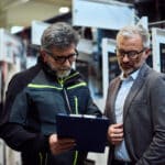 Ein Techniker und ein Vorgesetzter besprechen Unterlagen vor Schaltschränken in einer Industriehalle