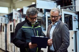 Zwei Männer blicken in einer Industrieanlage gemeinsam auf ein Klemmbrett