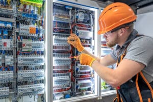 Elektriker mit Schutzhelm und Handschuhen arbeitet an einem komplexen Schaltschrank
